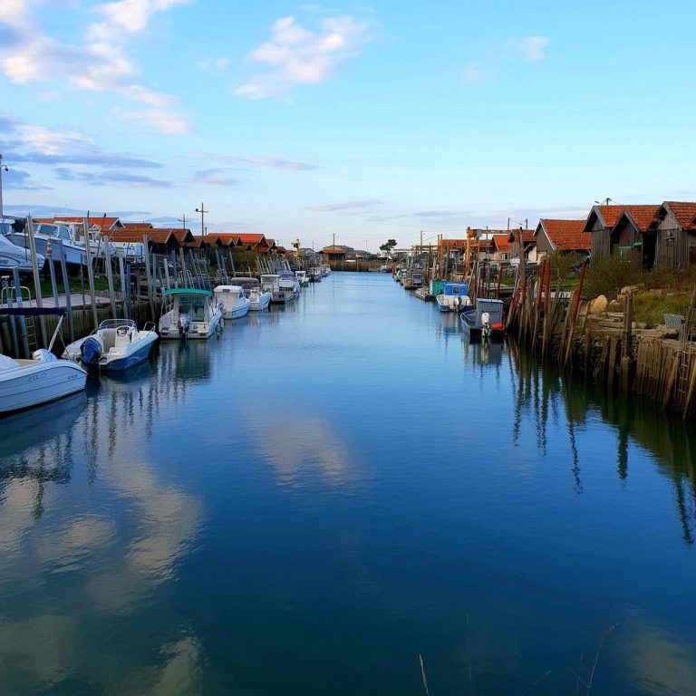 Port ostréicole paisible avec des bateaux, maisons colorées et ciel bleu.